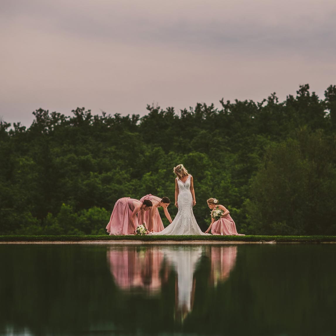 Bride in a white gown with bridesmaids in pink dresses adjusting her train by a reflective pool in Tuscany during a wedding in San Galgano Abbey