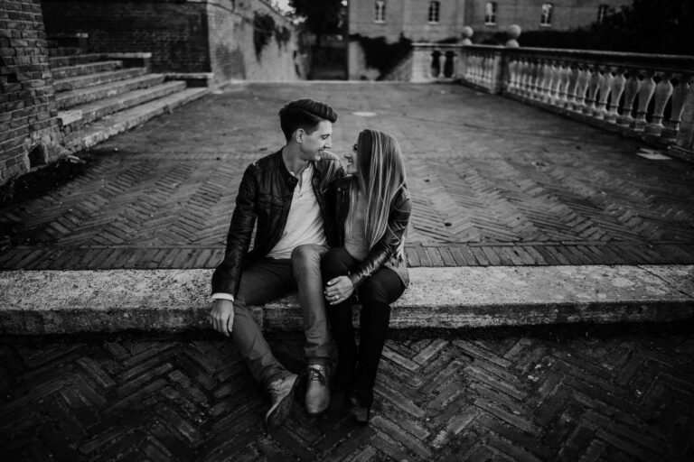 A couple sits close together on a stone ledge in an outdoor courtyard, gazing and smiling at each other. This Engagement and Portrait Couple moment is captured in black and white, with brick paving and historic buildings in the background. Tuscan Wedding Photographer