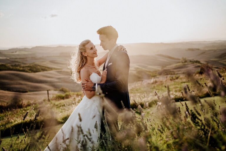 A bride and groom embrace in a sunlit field with rolling hills in the background, capturing the essence of their destination wedding in Tuscany. The brides white gown contrasts the grooms dark suit. Grasses and wildflowers surround them, bathed in the warm glow of the setting sun. Tuscan Wedding Photographer