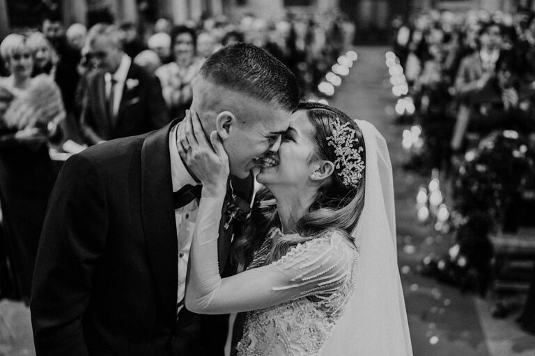 A black and white photo by Federico Pannacci captures a wedding ceremony where a couple shares a joyful moment. The bride, in a detailed gown with a veil and embellished headpiece, holds the grooms face as they lean in for a kiss during this luxury event for influencers and VIPs. Guests smile in the background. Tuscan Wedding Photographer