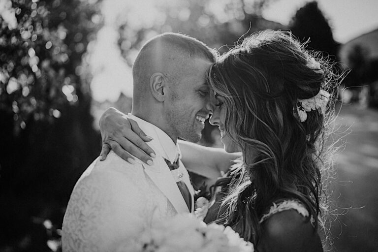 A black and white photo by Photos by Federico Pannacci captures a couple on their wedding day in an intimate embrace. The bride, with long wavy hair and floral accents, gently holds the groom in a light-colored suit. They touch foreheads, smiling amidst blurred trees and sunlight. Tuscan Wedding Photographer