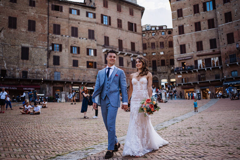 A bride in a lace wedding dress holds a bouquet, walking hand-in-hand with her groom in a light blue suit through a historic European square, both smiling—capturing the joy of their romantic wedding at Corsignano. Tuscan Wedding Photographer