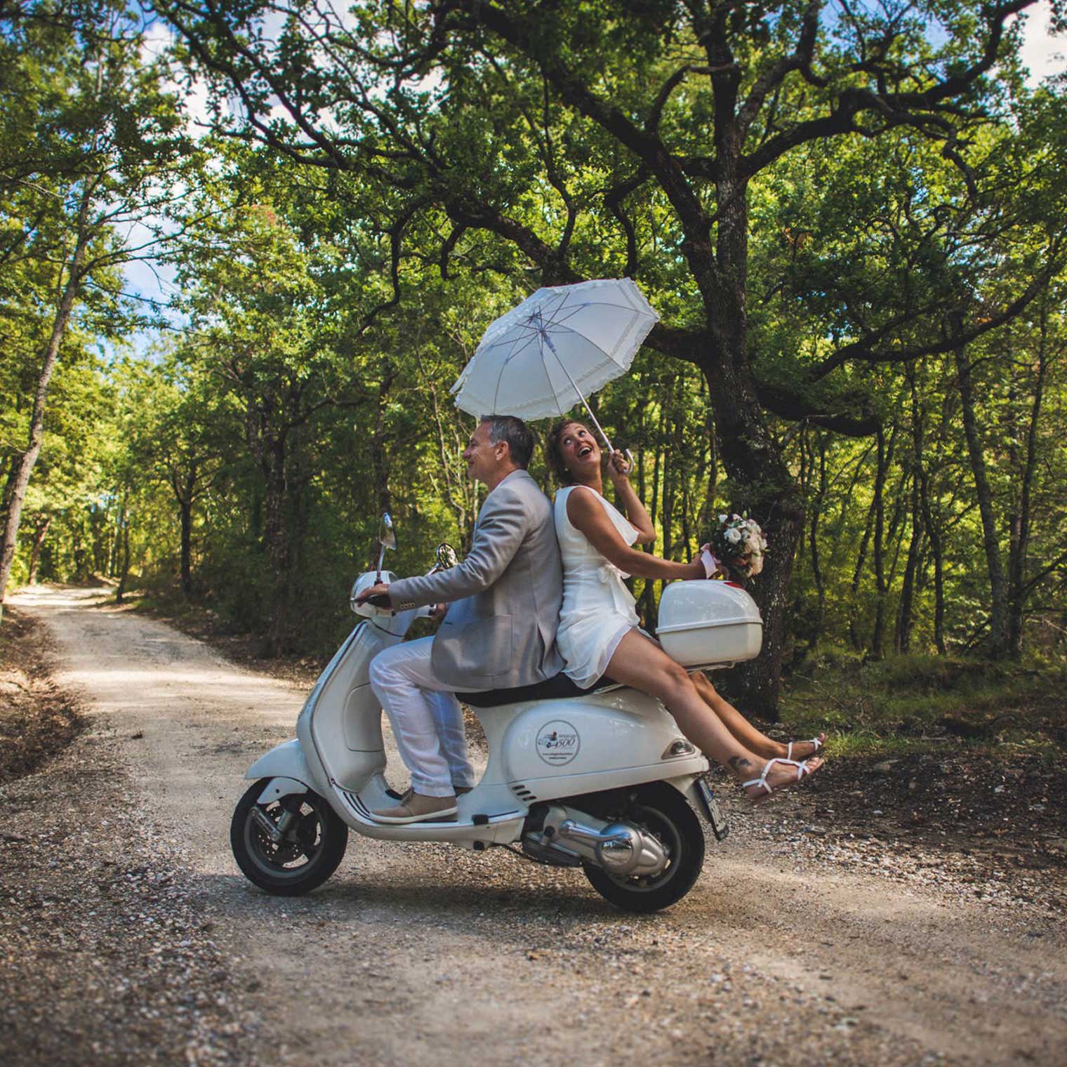 A couple in wedding attire sits on a vintage white scooter along a forest path. The groom, in a gray suit, drives while the bride, in a white dress, holds a parasol and bouquet, laughing. Captured by the best destination wedding photographer in Tuscany, sunlight filters through the trees in this romantic scene. Tuscan Wedding Photographer
