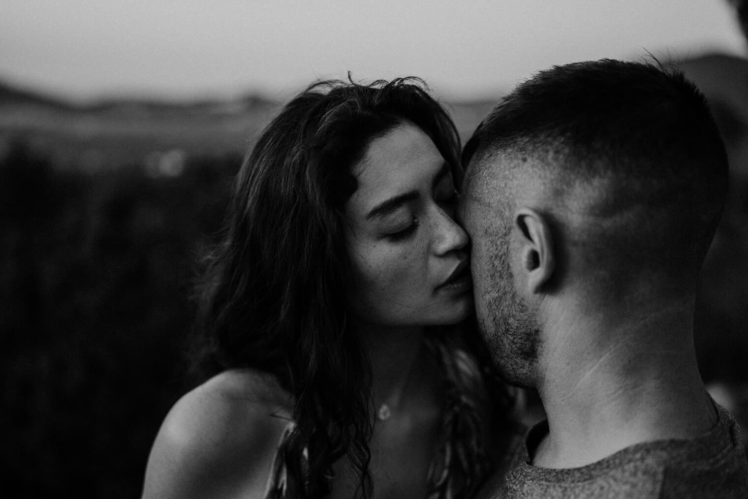 A black-and-white photo captures a tender, intimate moment as a woman with long hair leans in to kiss a man on the cheek. The man, with short hair and a trimmed beard, stands outdoors against blurred Tuscan hills, embodying the artistry of the Best Destination Wedding Photographer in Tuscany. Tuscan Wedding Photographer
