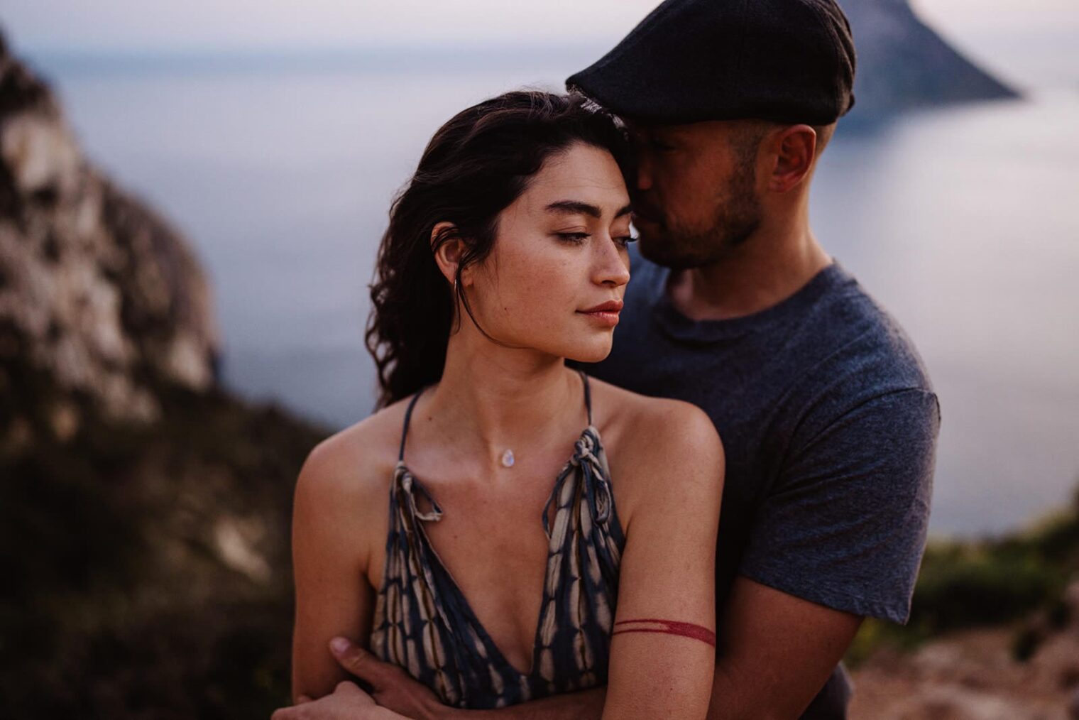A couple stands on a cliffside, overlooking the ocean. The woman, with long dark hair and a patterned dress, leans affectionately against the man, who wears a cap and a gray shirt. Captured by the best destination wedding photographer in Tuscany, they share an intimate moment as calm waters shimmer at dusk. Tuscan Wedding Photographer