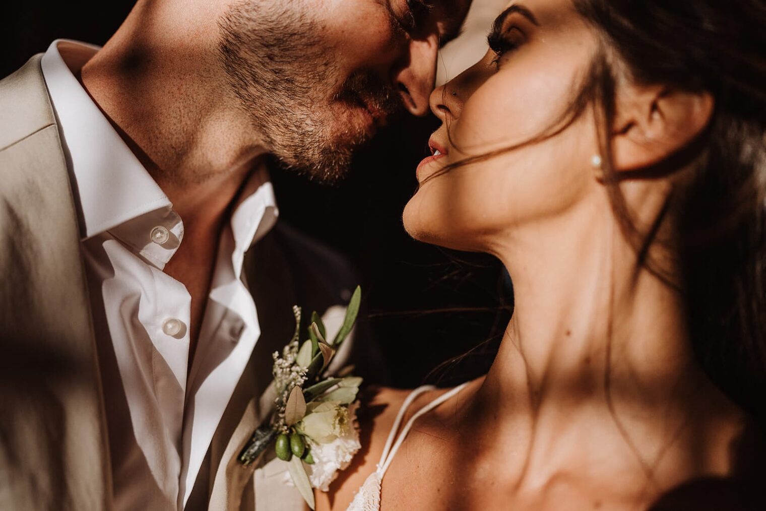 Close-up of a couple in an intimate moment, captured by the best destination wedding photographer in Tuscany. With their faces almost touching, the man wears a light suit with a white shirt and boutonniere, while the woman’s long hair and pearl earrings are softly highlighted by warm lighting, creating a romantic atmosphere. Tuscan Wedding Photographer