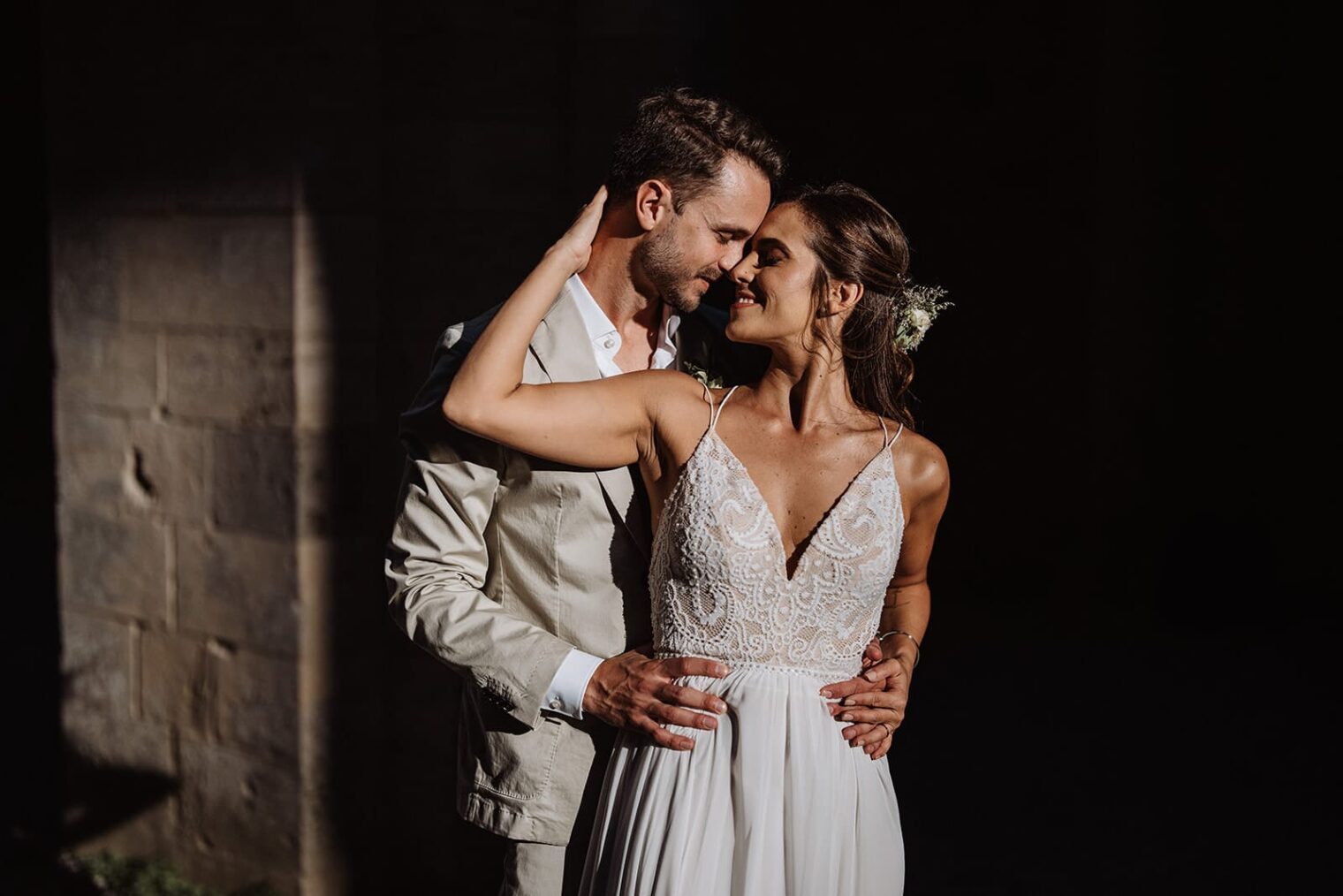 A couple in wedding attire stands closely, embraced and smiling at each other. The bride wears a white, lace-embellished gown, and the groom dons a light beige suit. Captured by the best destination wedding photographer in Tuscany, sunlight casts shadows that highlight their joy against a dark, textured wall. Tuscan Wedding Photographer