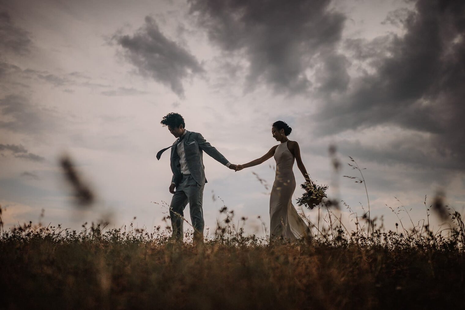 A couple walks hand in hand across a field under a dramatic, cloudy sky, captured by the best destination wedding photographer in Tuscany. The man wears a suit while the woman is in a flowing dress, holding a bouquet. Tall grasses sway around them, creating a romantic and serene atmosphere. Tuscan Wedding Photographer