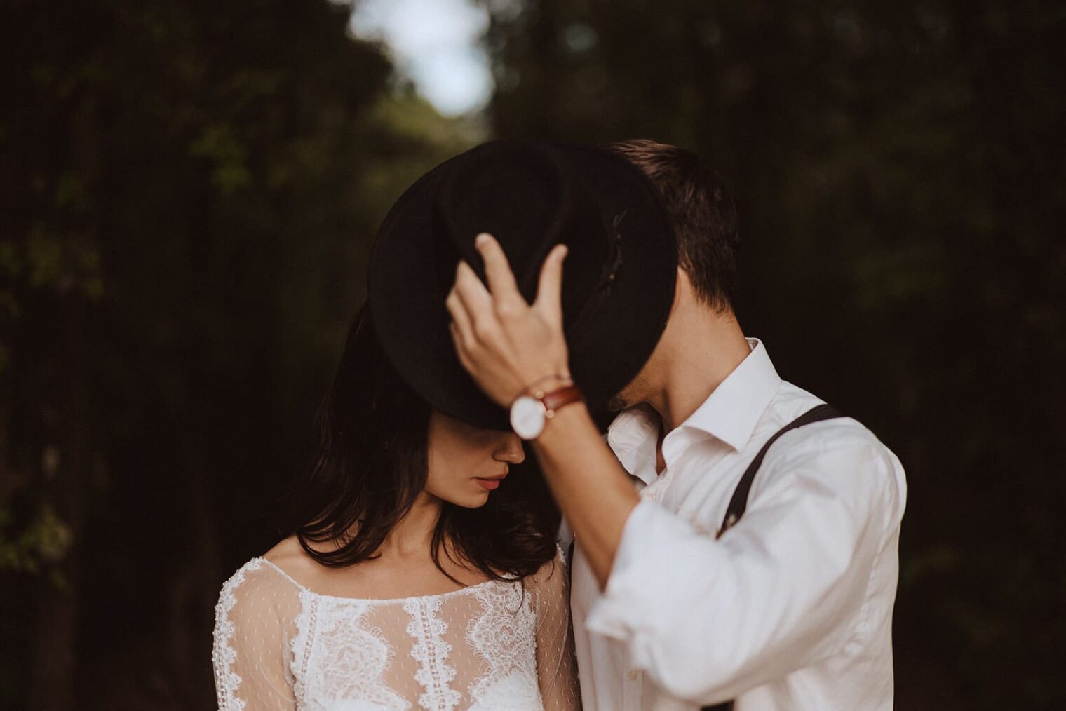 A couple stands closely in a forest setting, evoking the charm captured by the best destination wedding photographer in Tuscany. The man, in a white shirt and suspenders, covers both their faces with a black hat. The woman, wearing a lace dress, gazes downwards amidst blurred trees creating an intimate ambiance. Tuscan Wedding Photographer