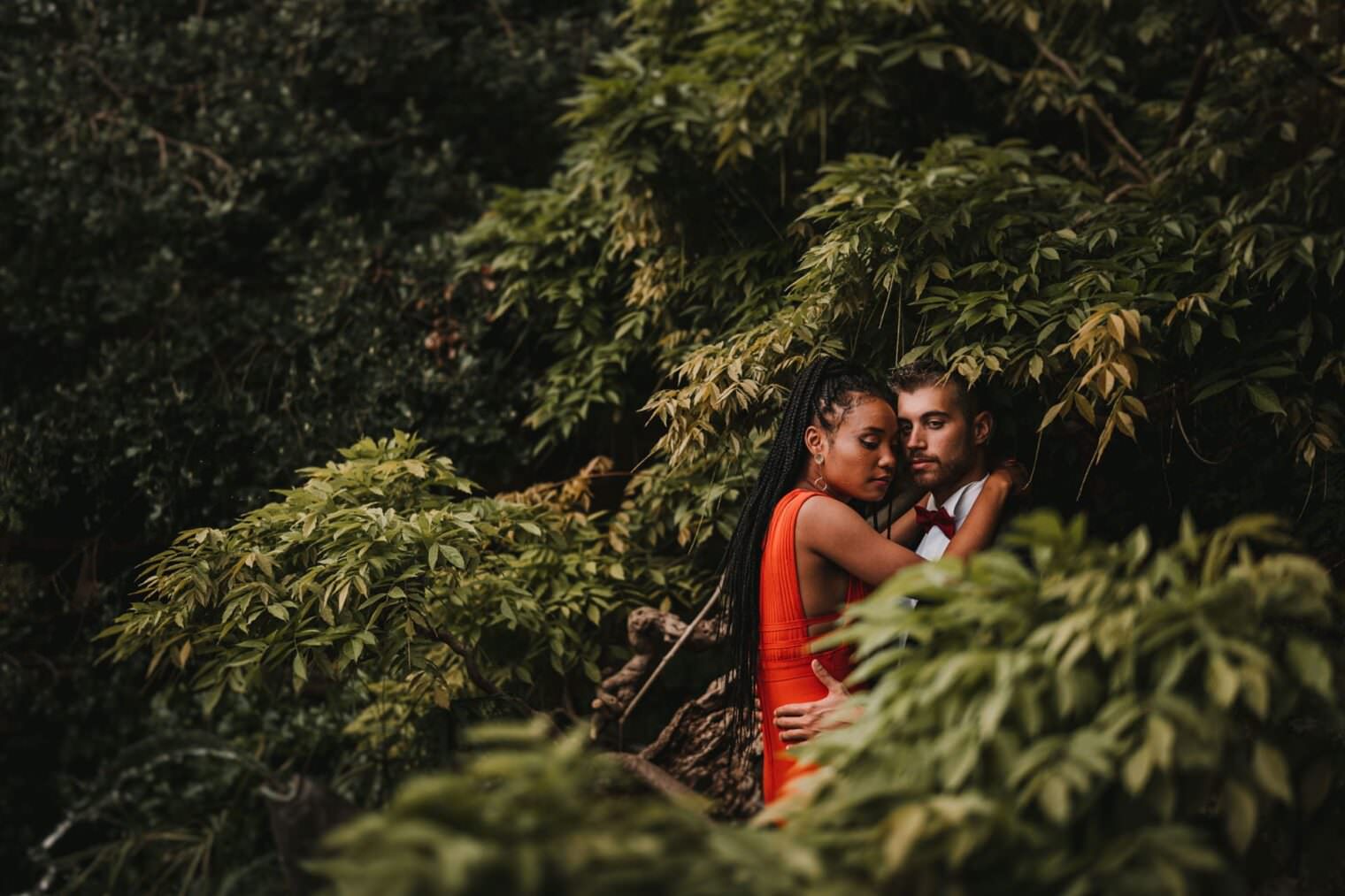 A couple embraces amid lush greenery. The woman in a sleeveless red dress rests her head on the mans shoulder while he gazes ahead thoughtfully, creating a serene, intimate atmosphere. Captured by the best destination wedding photographer in Tuscany, their love story unfolds amidst the dense foliage. Tuscan Wedding Photographer