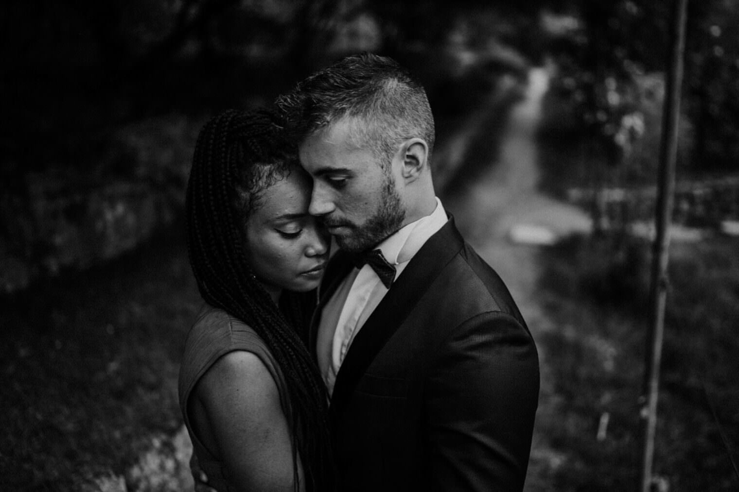 A black and white photo of a couple embracing closely, heads touching, eyes closed portrays an intimate moment captured by the best destination wedding photographer in Tuscany. The man wears a suit and the woman, with braided hair in a sleeveless top, stand on a tree-lined path evoking serenity. Tuscan Wedding Photographer
