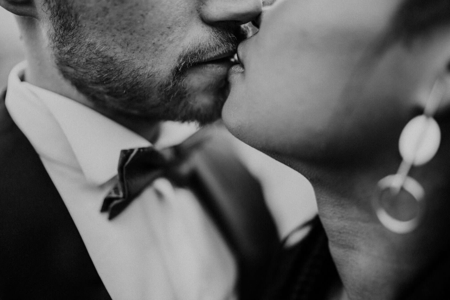 Close-up black and white image of two people kissing, taken by the best destination wedding photographer in Tuscany. The scene captures the lower half of their faces, focusing on the intimate moment. The person on the left has a beard and wears a bow tie; the person on the right has earrings. Tuscan Wedding Photographer