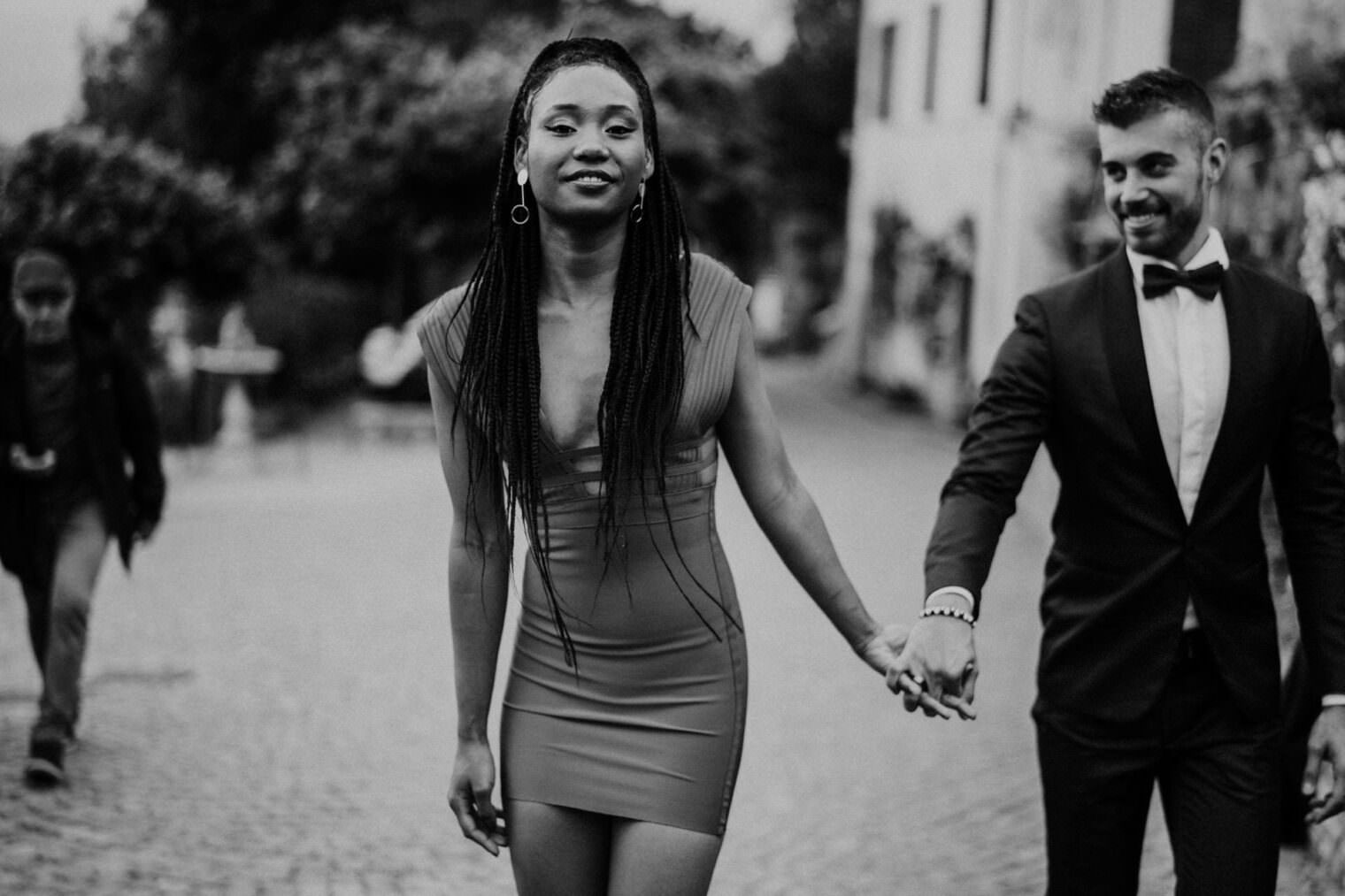 A black and white photo captures a smiling woman with braided hair in a form-fitting dress walking hand in hand with a bearded man in a suit and bow tie. On the cobblestone path framed by blurred trees and buildings, this image is worthy of the best destination wedding photographer in Tuscany. Tuscan Wedding Photographer