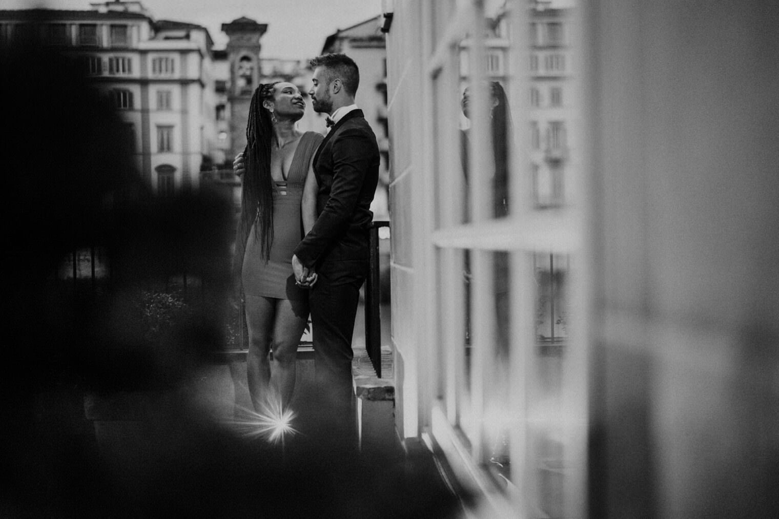 A black and white photo captures a couple on an outdoor terrace. The woman in a dress holds the mans hand as they gaze at each other, embodying the magic promised by the best destination wedding photographer in Tuscany. A building with arched windows stands behind, and a lens flare adds a starburst effect below. Tuscan Wedding Photographer