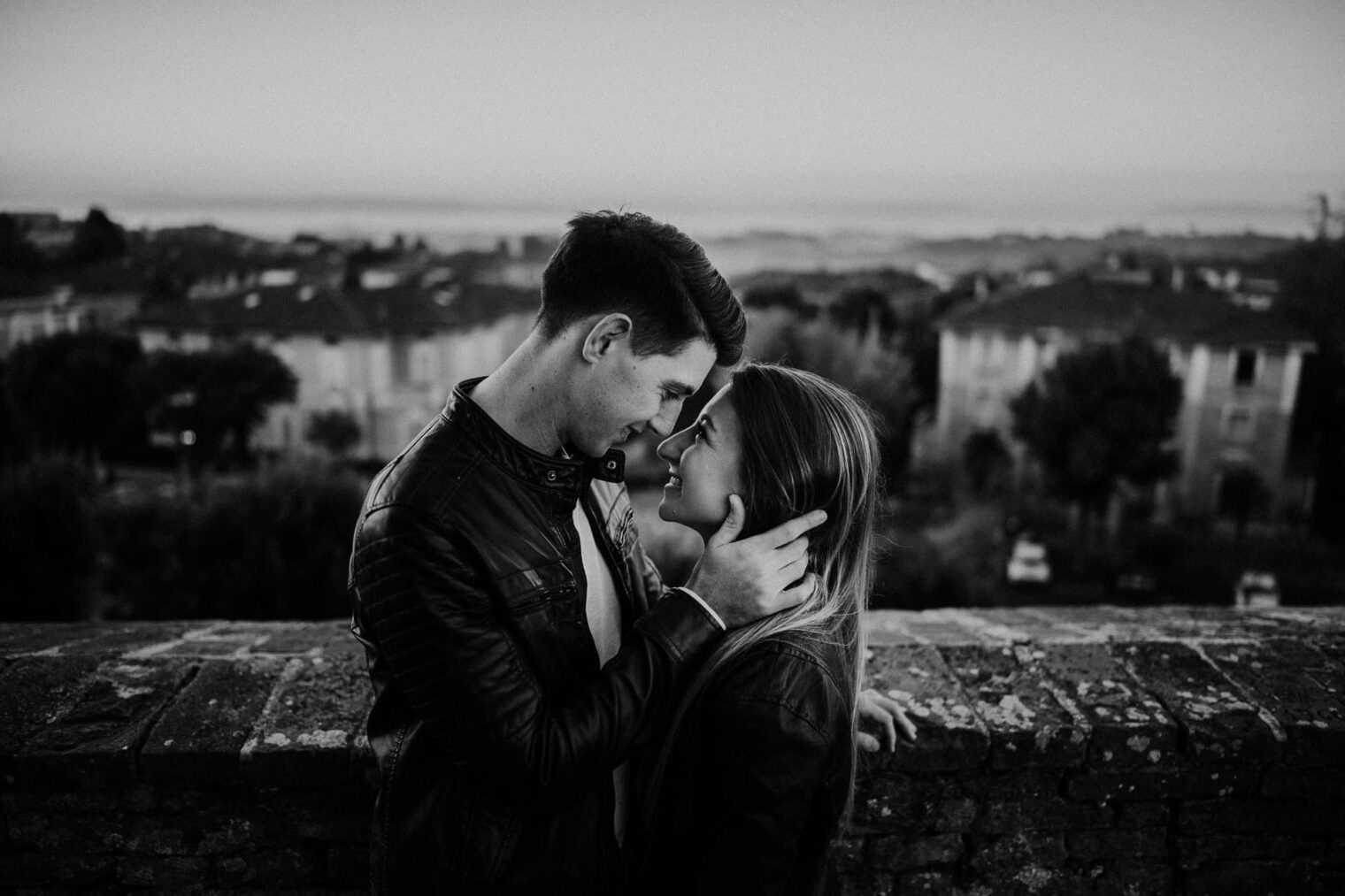 A couple stands close, gazing into each others eyes against a brick wall. The man gently holds the woman’s face, both smiling softly in their leather jackets. With rooftops and trees under a dimly lit sky, this black-and-white scene captures a moment worthy of the Best Destination Wedding Photographer in Tuscany. Tuscan Wedding Photographer