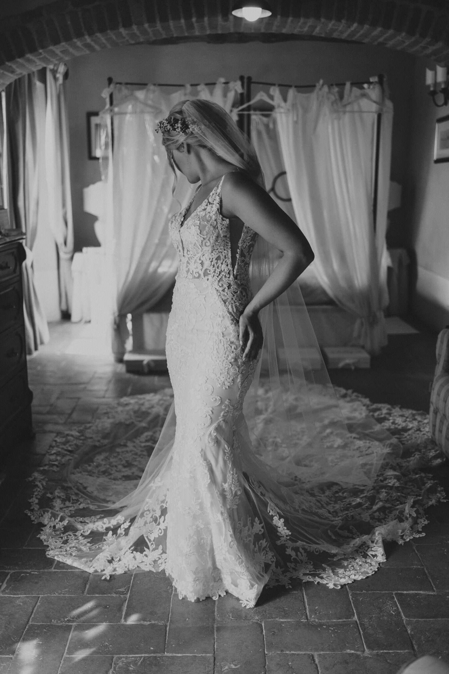 A bride in an intricately designed lace wedding gown stands in a dimly lit room with rustic brick flooring. Captured by the best destination wedding photographer in Tuscany, her dress features a long train and a sheer veil. The romantic setting includes floral hair accessories, a canopy bed, and heavy curtains. Tuscan Wedding Photographer