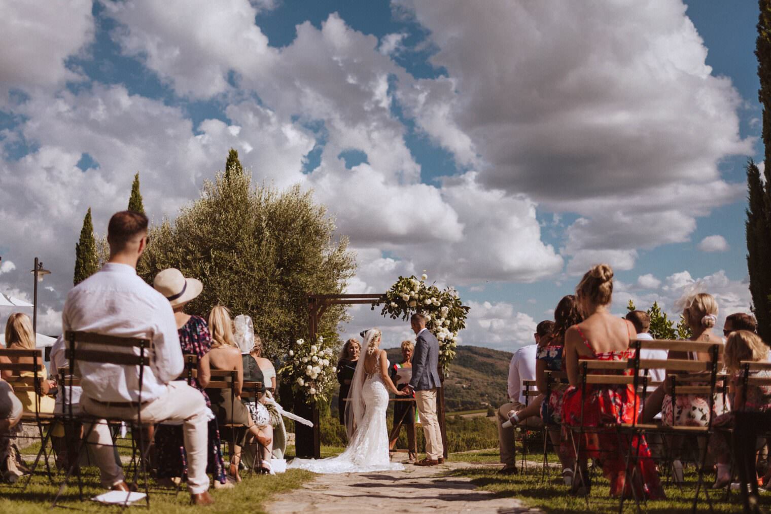 A couple stands under a floral arch exchanging vows at an outdoor wedding ceremony, artfully captured by the best destination wedding photographer in Tuscany. Guests are seated on wooden chairs amid lush greenery, with a scenic view and fluffy clouds painting a picturesque atmosphere. Tuscan Wedding Photographer