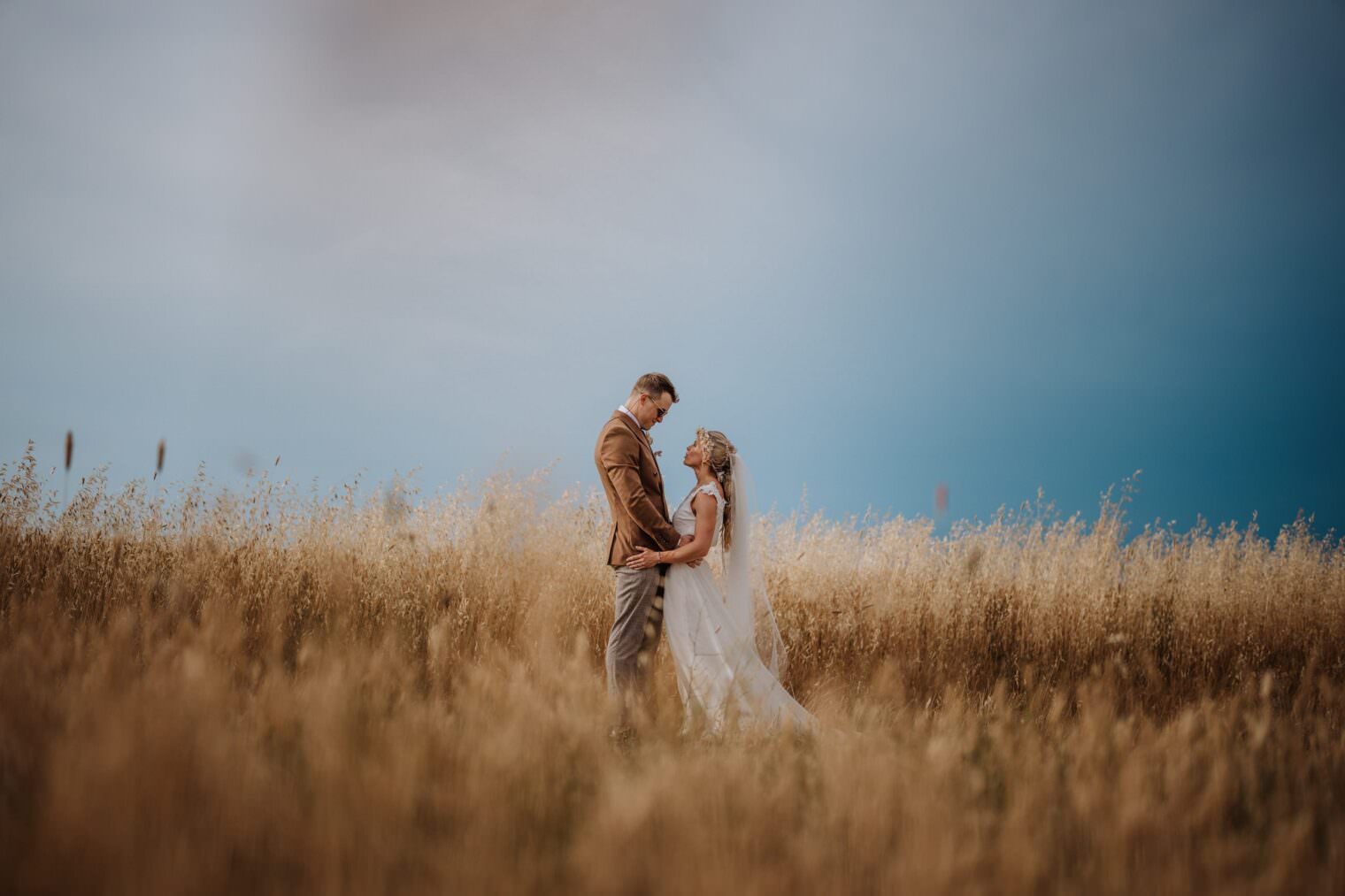 A couple stands in a field of tall, golden grass under a cloudy sky. The man in a brown jacket and gray pants holds hands with the woman in a flowing white dress and veil. Captured by the best destination wedding photographer in Tuscany, they embrace amidst the vast open landscape. Tuscan Wedding Photographer