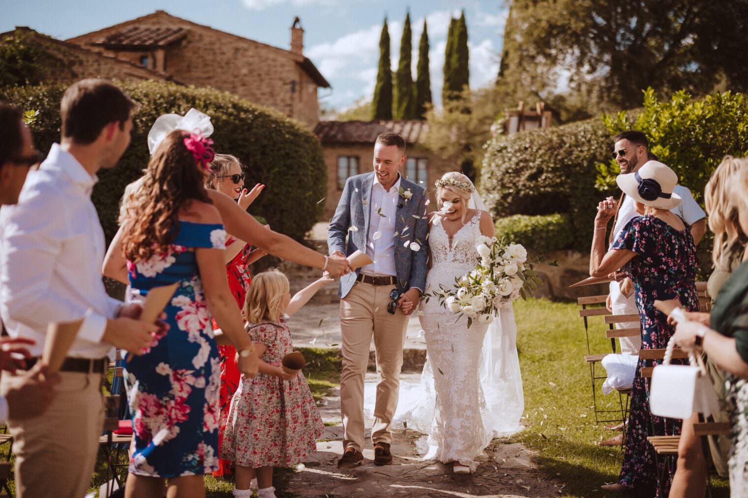 A bride and groom, captured by the best destination wedding photographer in Tuscany, walk down an outdoor aisle, smiling. The brides white lace dress and veil complement her bouquet. The groom sports a gray blazer with beige pants. Guests cheer and toss confetti in a sunny garden beside a rustic building. Tuscan Wedding Photographer
