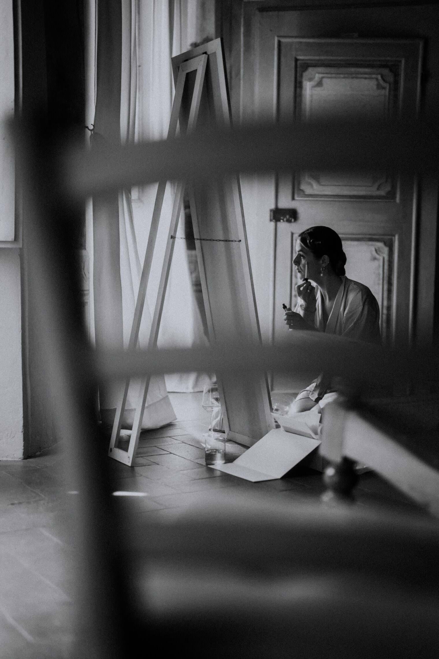 A black and white photo captures a person in a robe sitting on the floor, applying makeup before a large mirror. In a room with wooden floors and sheer-curtained windows, the scene is beautifully framed by a chair in the foreground—a moment elegantly captured by the Best Destination Wedding Photographer in Tuscany. Tuscan Wedding Photographer