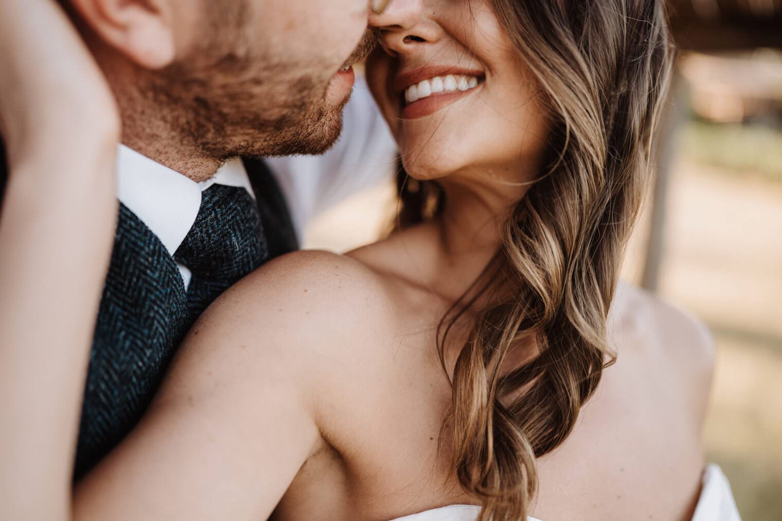 Close-up of a couple embracing and smiling. The woman, in a strapless white dress, has long, wavy brown hair. The man sports a beard and wears a white shirt with a dark textured tie and suit vest. Captured by the best destination wedding photographer in Tuscany, they are outdoors, bathed in warm light. Tuscan Wedding Photographer