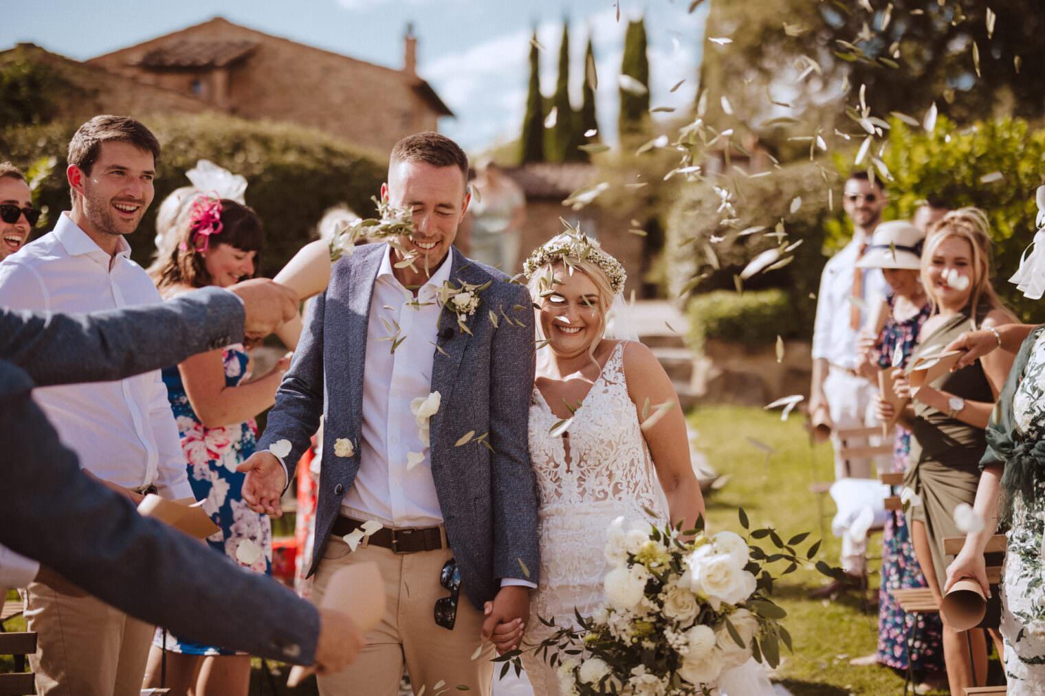 A bride and groom walk joyfully hand in hand down an aisle, surrounded by guests throwing flower petals. The scene is beautifully captured by the best destination wedding photographer in Tuscany. The brides lace dress complements her floral crown, while the groom dons a gray blazer amidst vibrant summer attire and lush greenery. Tuscan Wedding Photographer
