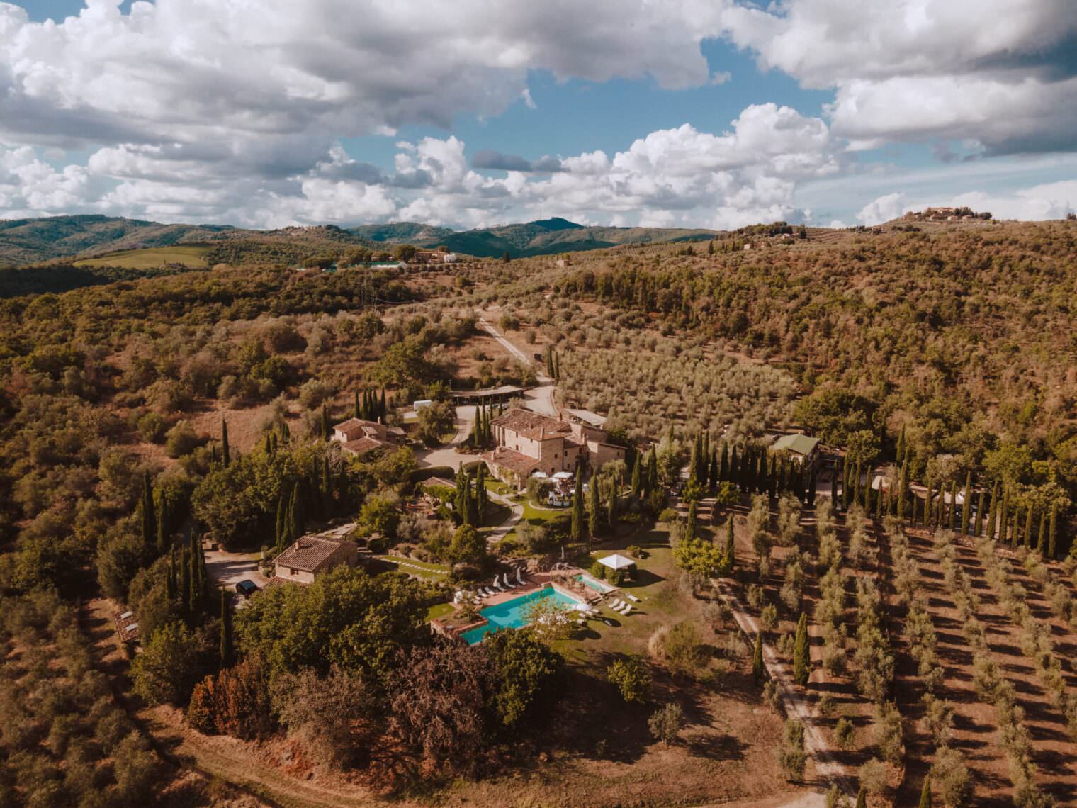 Aerial view of a picturesque countryside estate in Tuscany, perfect for the best destination wedding. Featuring a central rustic villa surrounded by lush greenery, with a clear blue swimming pool and loungers. Rolling hills and a cloudy sky complete this stunning backdrop. Tuscan Wedding Photographer