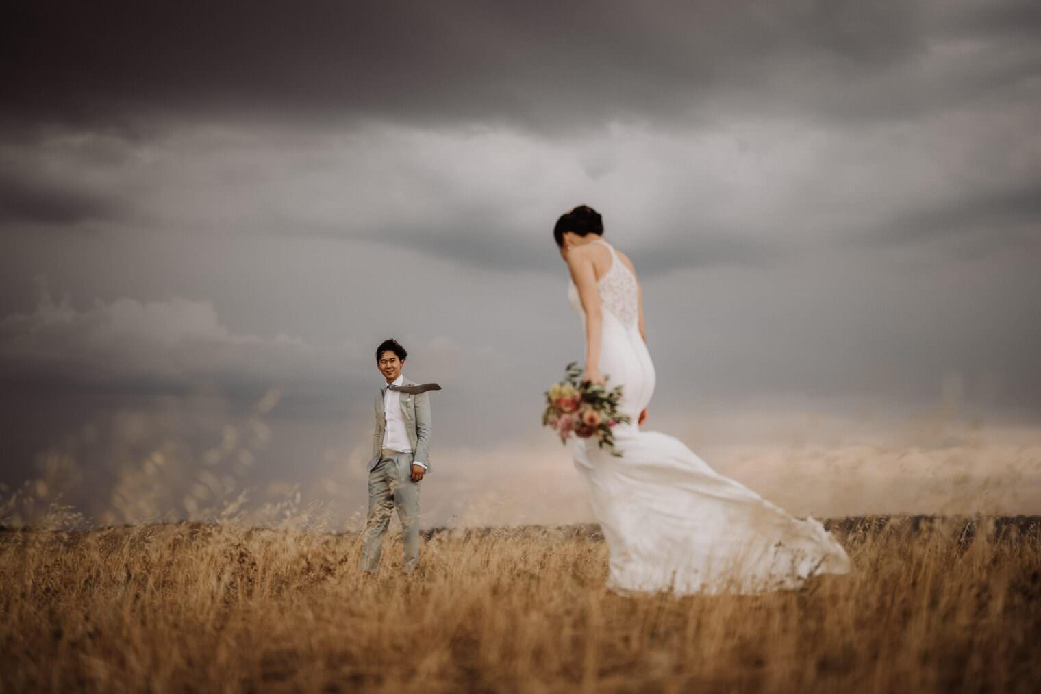 A bride in a flowing white dress holds a bouquet, walking through a field of tall, golden grass. As the groom in a light gray suit smiles from the background, the best destination wedding photographer in Tuscany captures the drama of overcast skies and moody clouds surrounding this enchanting scene. Tuscan Wedding Photographer