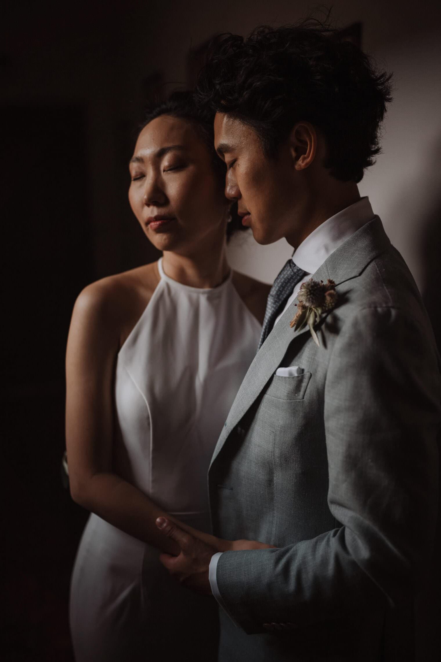 A couple embraces tenderly in dim lighting, captured by the best destination wedding photographer in Tuscany. The woman, eyes closed, dons a white halter-neck dress. The man wears a light gray suit with a floral boutonniere and checkered tie. They hold hands, conveying intimacy and calmness. Tuscan Wedding Photographer