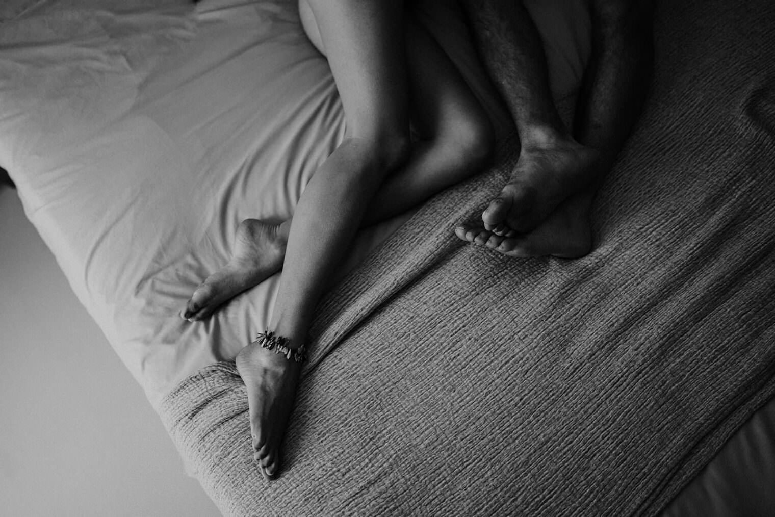 A black and white image captures an intimate moment as two people lie on a bed, their legs intertwined under a light sheet and textured blanket. An anklet adorns one persons ankle, echoing the delicate artistry youd expect from the best destination wedding photographer in Tuscany. Tuscan Wedding Photographer