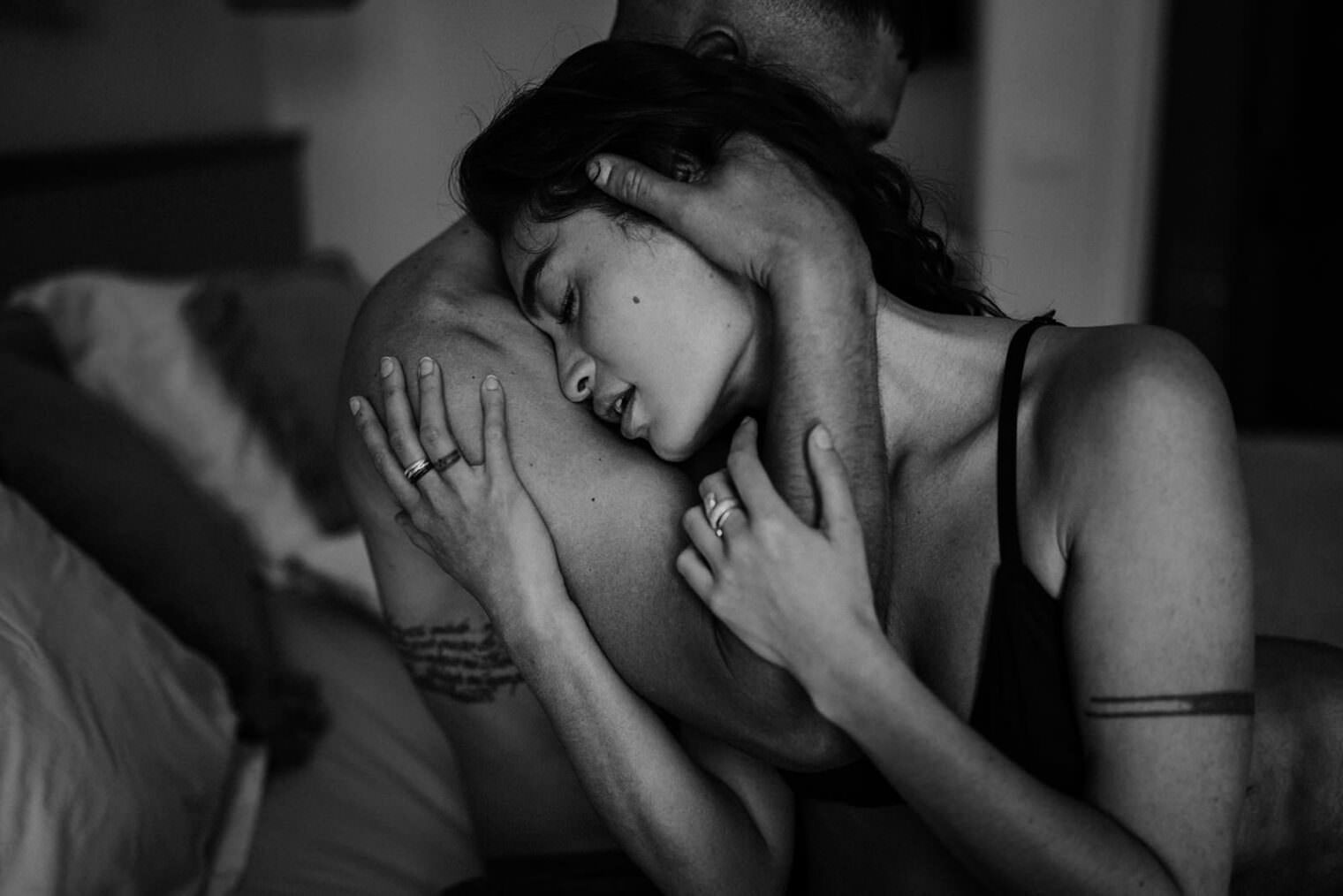 A black and white photo captures a couple embracing intimately, reminiscent of a romantic scene youd expect from the best destination wedding photographer in Tuscany. The woman, in a bra, rests her head on the mans bare shoulder, her serene expression complementing the way her hands caress his neck and back adorned with rings. Tuscan Wedding Photographer