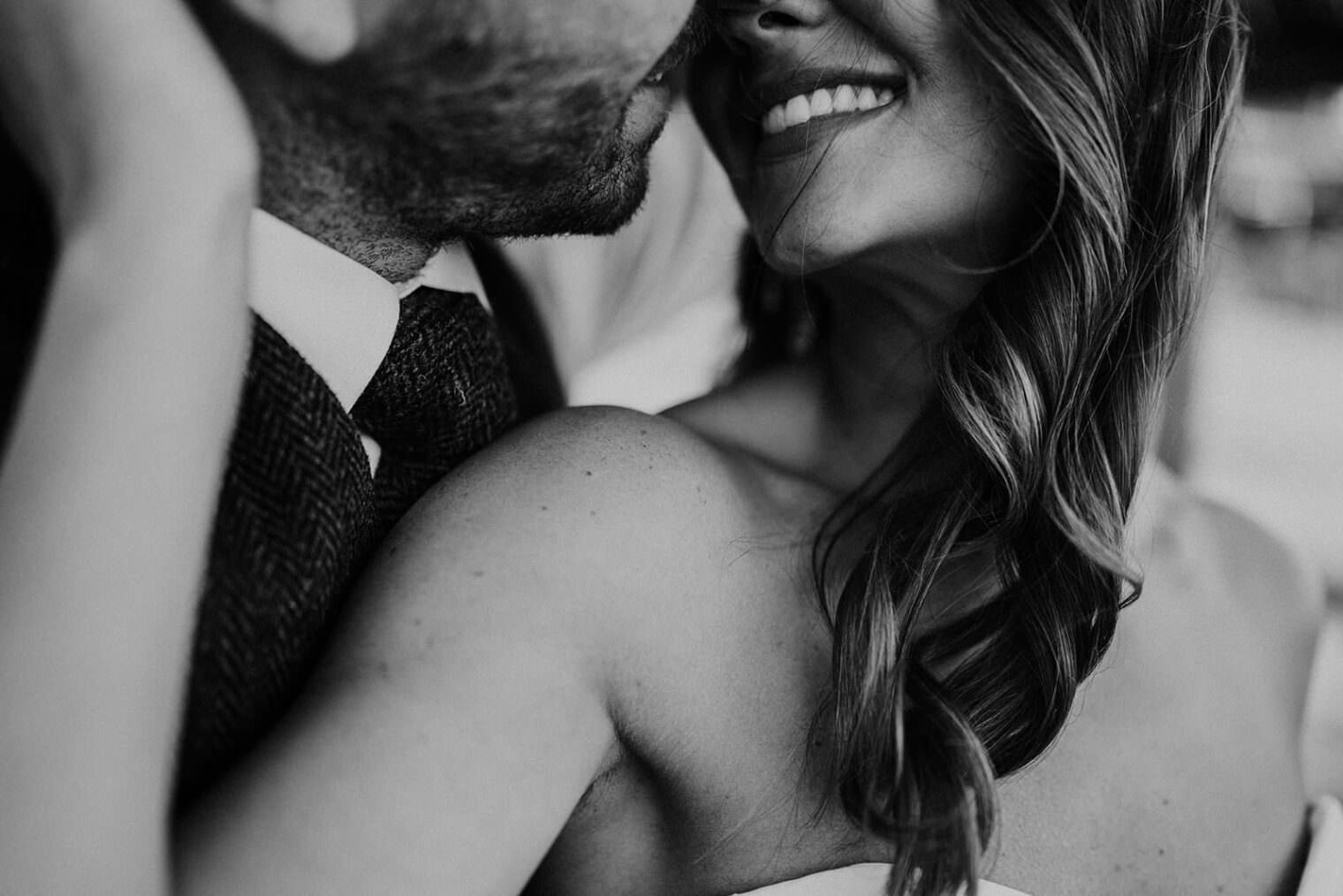A black and white close-up of a couple embracing captures the essence of love. The woman, smiling beneath wavy hair and a strapless top, leans into her suited partner with a textured tie. Their intertwined arms embody romance, much like those cherished by the best destination wedding photographer in Tuscany. Tuscan Wedding Photographer