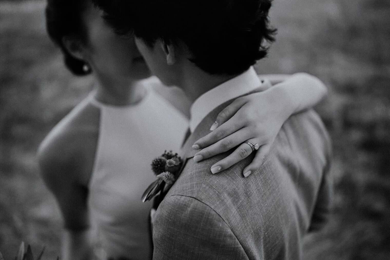 A grayscale photo captures a couple closely embracing, with the woman’s engagement ring shimmering subtly. Her sleeveless dress complements his suit adorned with a boutonniere. Their partially obscured faces enhance the intimate mood, echoing the artistry of the best destination wedding photographer in Tuscany. Tuscan Wedding Photographer