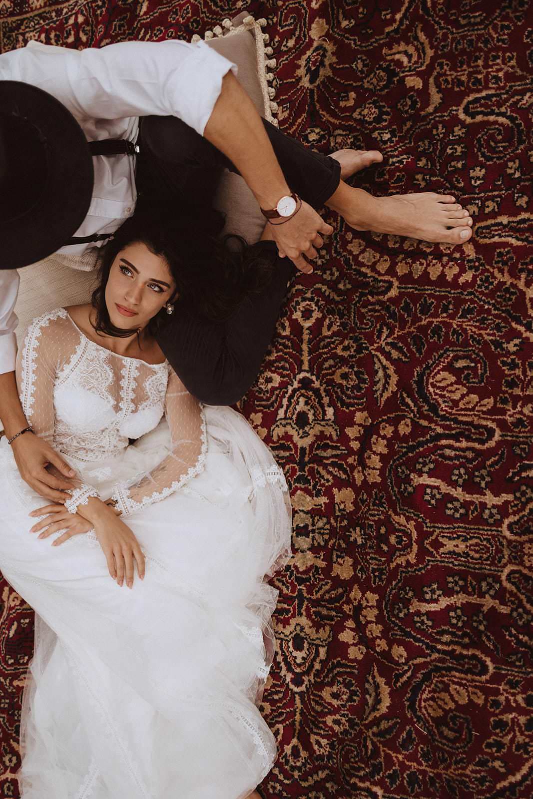 A couple relaxes on an ornate maroon and gold patterned rug. The woman, in a white lace wedding dress, rests her head on the mans lap. Hes in a white shirt and black pants, his arm around her with his watch visible. Captured by the best destination wedding photographer in Tuscany, bare feet are shown. Tuscan Wedding Photographer
