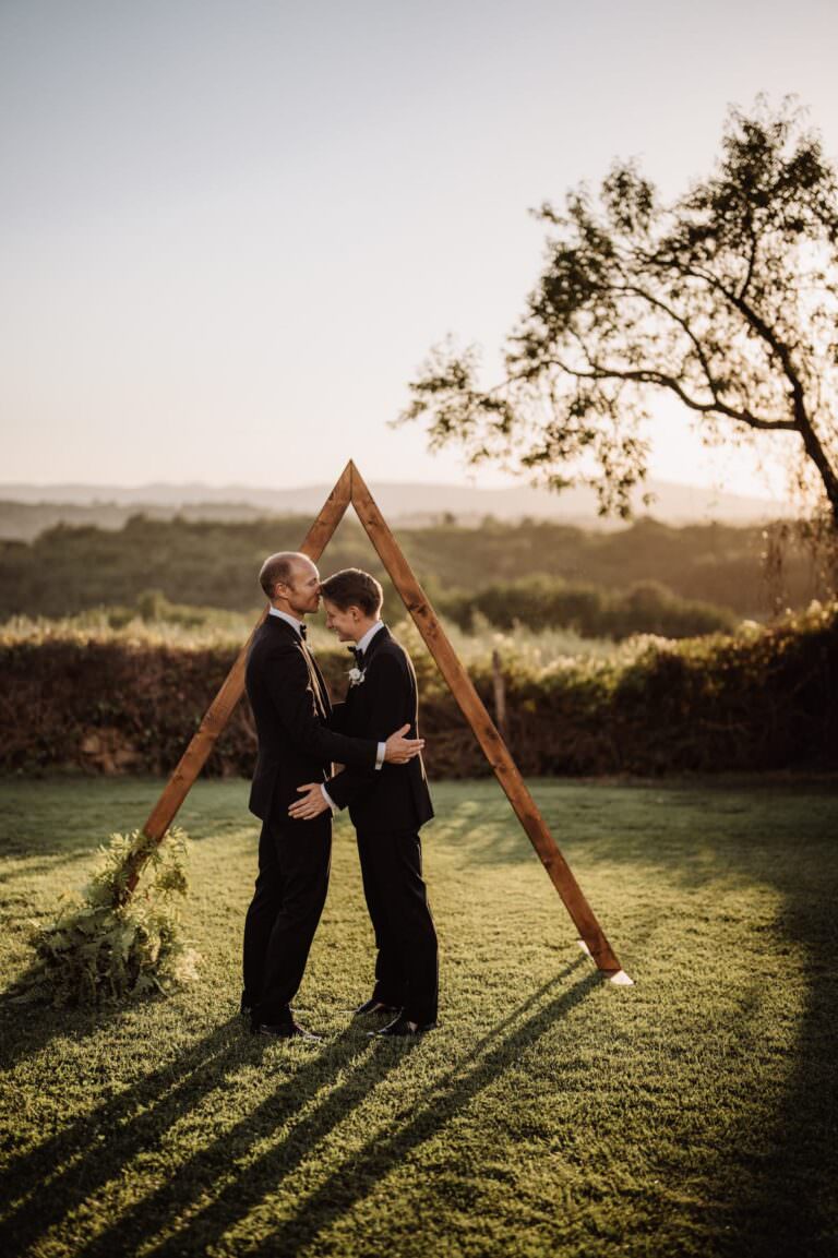 Two people in suits stand facing each other, holding hands under a wooden triangular arch at Certosa di Pontignano in Tuscany. The lush landscape with distant hills and a tree to the right enhances the serene and romantic atmosphere of this beautiful same-sex wedding at sunset. Tuscan Wedding Photographer