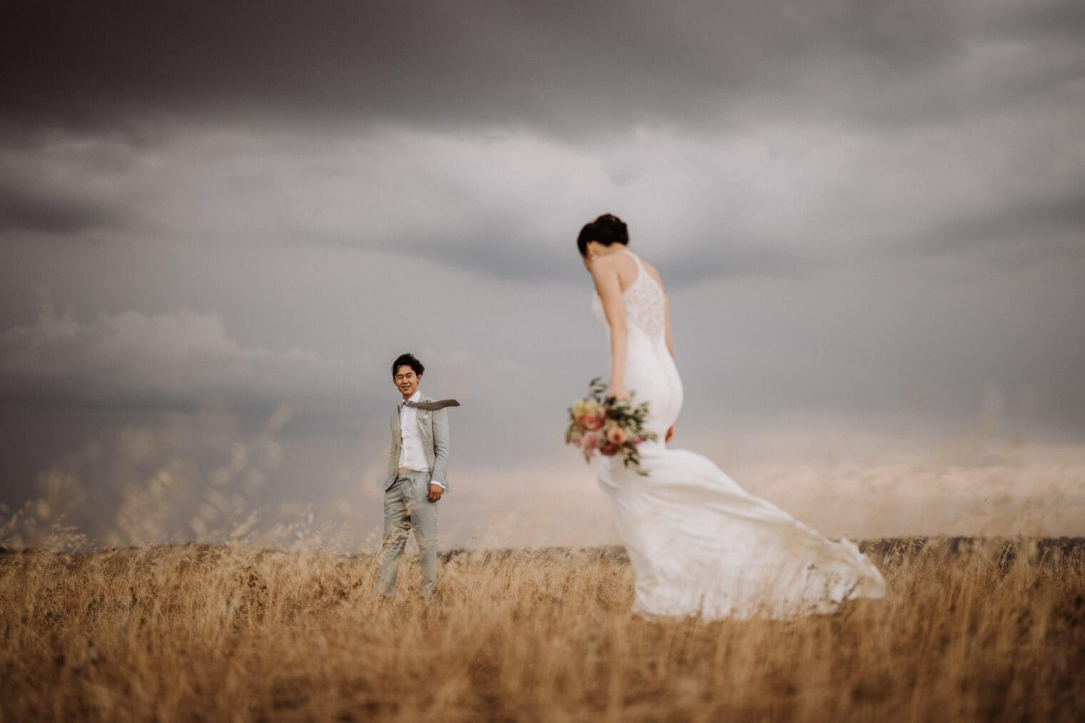 A bride in a flowing white dress holds a bouquet as she walks towards a groom in a gray suit. They stand in the golden fields of Val dOrcia in Tuscany under a cloudy sky, creating a romantic and atmospheric scene reminiscent of a wedding at il Ringo. The groom smiles as he watches her approach. Tuscan Wedding Photographer