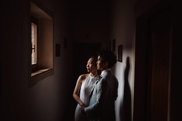 A couple stands in a dimly lit hallway at il Ringo, with soft light streaming through a small window. The woman, in a sleeveless white dress, and the man, in a light gray suit, gaze thoughtfully outside. Framed pictures hang on the dark walls, creating an intimate and serene atmosphere reminiscent of Val dOrcia in Tuscany. Tuscan Wedding Photographer