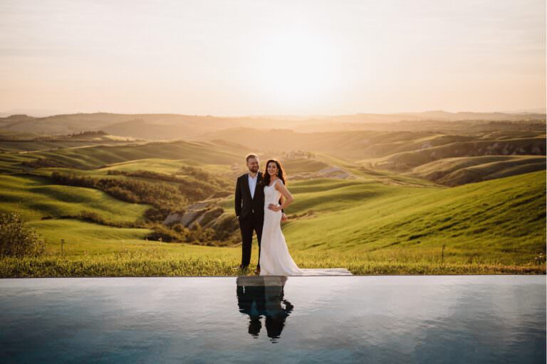 A couple in formal attire stands by a reflective pool at sunset during their British wedding at Lazy Olive. With Tuscanys rolling green hills in the background, the man wears a black suit, and the woman wears a white gown. Federico Pannacci captures this serene and romantic moment in the golden light. Tuscan Wedding Photographer