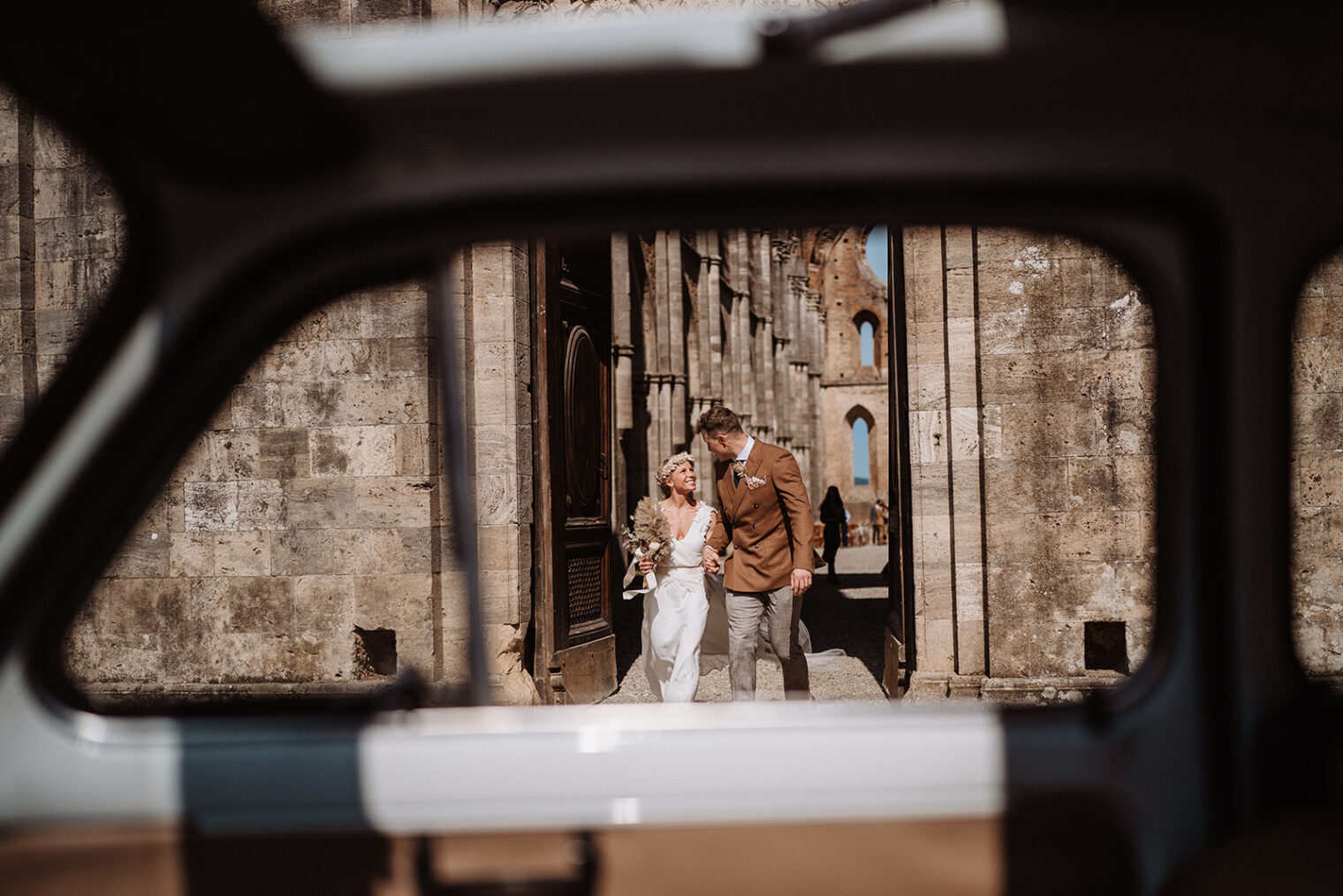 The photo captures a couple walking outside the stunning San Galgano Abbey, with the view being captured through the glass of an old 500 car. The couple is walking hand in hand, the man is wearing a brown suit and the woman is wearing a white wedding dress. They look happy and in love, deep in conversation as they walk. The abbey is visible in the background, its ancient stone walls and Gothic architecture providing a striking contrast to the couple's modern attire. The sun is shining down on the scene, casting a warm glow over the couple and highlighting the intricate details of the abbey's architecture. The car window provides a frame to the picture, giving the sense of a vintage and romantic atmosphere. The use of the car window also gives a sense of movement as if they are on a journey together. Overall the photo conveys a sense of love and history, capturing a special moment in a beautiful and historic setting.