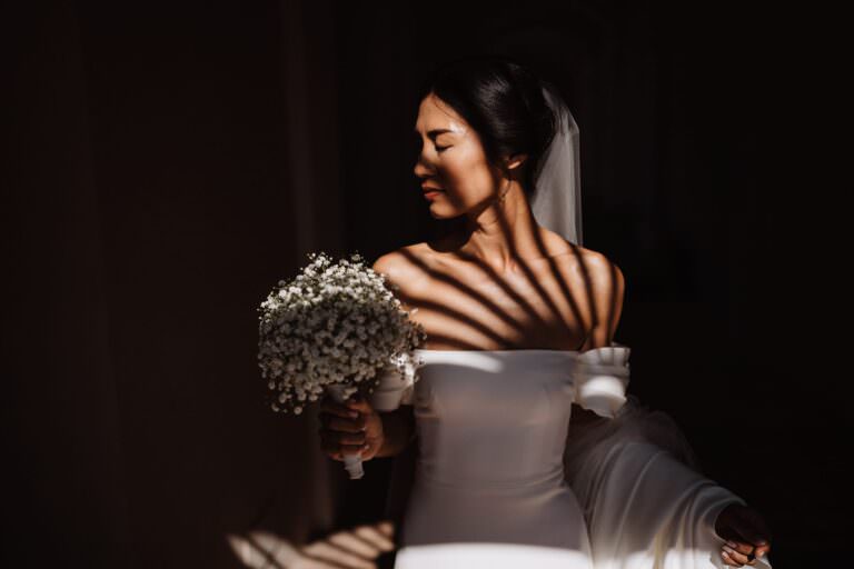 Against the dramatic lighting of Tropea, a bride in a strapless white gown holds a bouquet of white flowers. The striped shadows create an artistic touch on her serene face and dress. Her dark hair is styled up with a veil, capturing the essence of destination wedding photography in South Italy. Tuscan Wedding Photographer