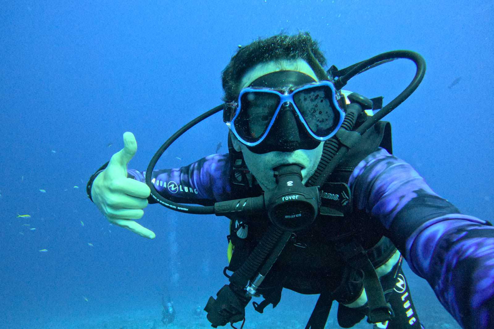 A scuba diver in a purple patterned wetsuit poses underwater, making a shaka hand gesture. They channel the adventurous spirit of the life of Federico Pannacci, framed by a black mask and breathing regulator. Bubbles rise around as small fish complete the scene in the deep blue ocean. Tuscan Wedding Photographer