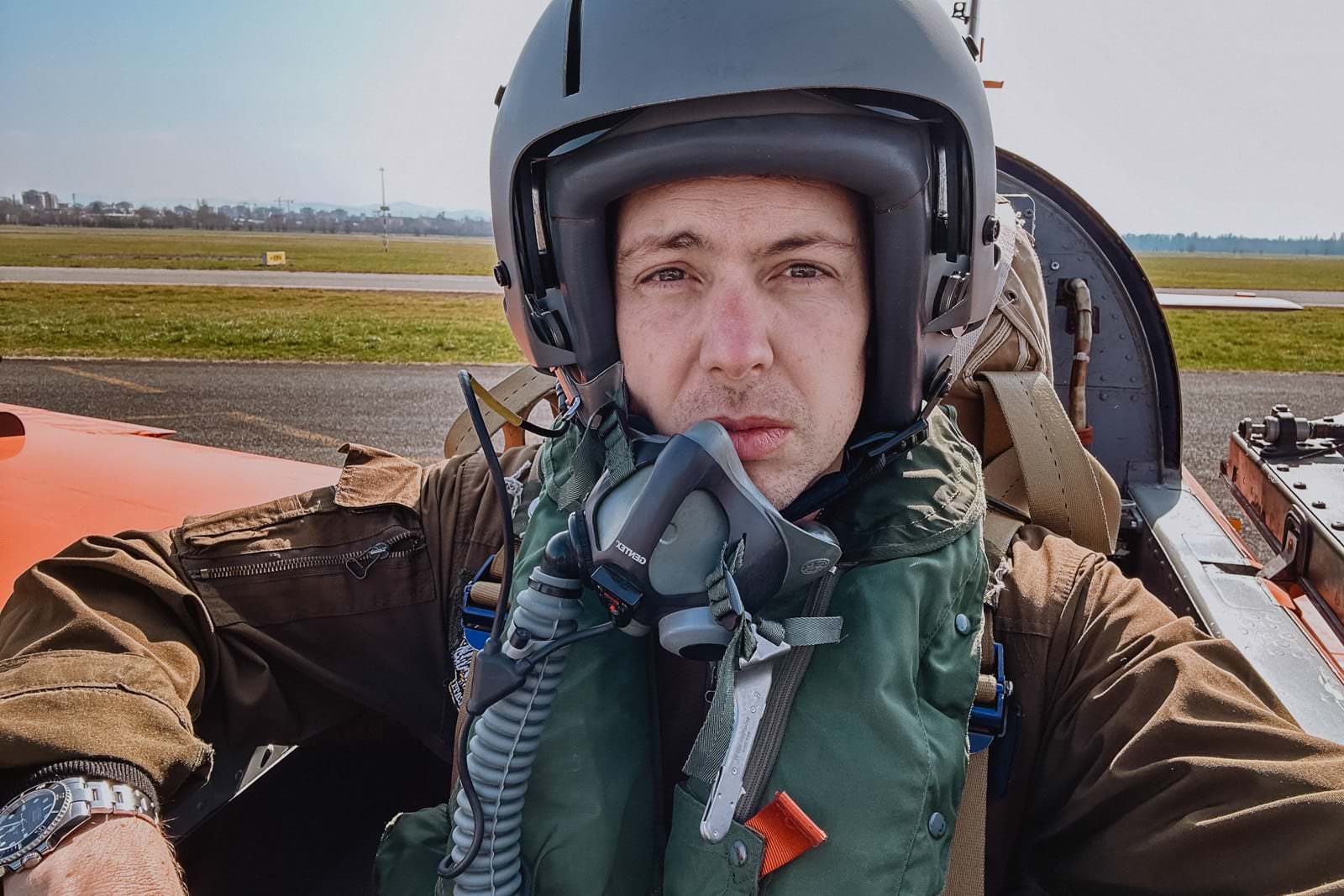 A person wearing a flight helmet and oxygen mask sits in the cockpit of an aircraft, reminiscent of a wedding photographer in Tuscany capturing breathtaking aerial shots. They are dressed in a brown jacket and green flight vest, with the runway and grassy area visible beneath the clear Italian sky. Tuscan Wedding Photographer