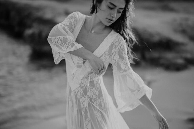 A woman in a flowing lace dress dances gracefully on a beach, her eyes closed and hair softly blown by the breeze. Captured by a Balearic wedding photographer, the black-and-white image enhances the ethereal atmosphere, focusing on her expressive movement and intricate dress details. Tuscan Wedding Photographer