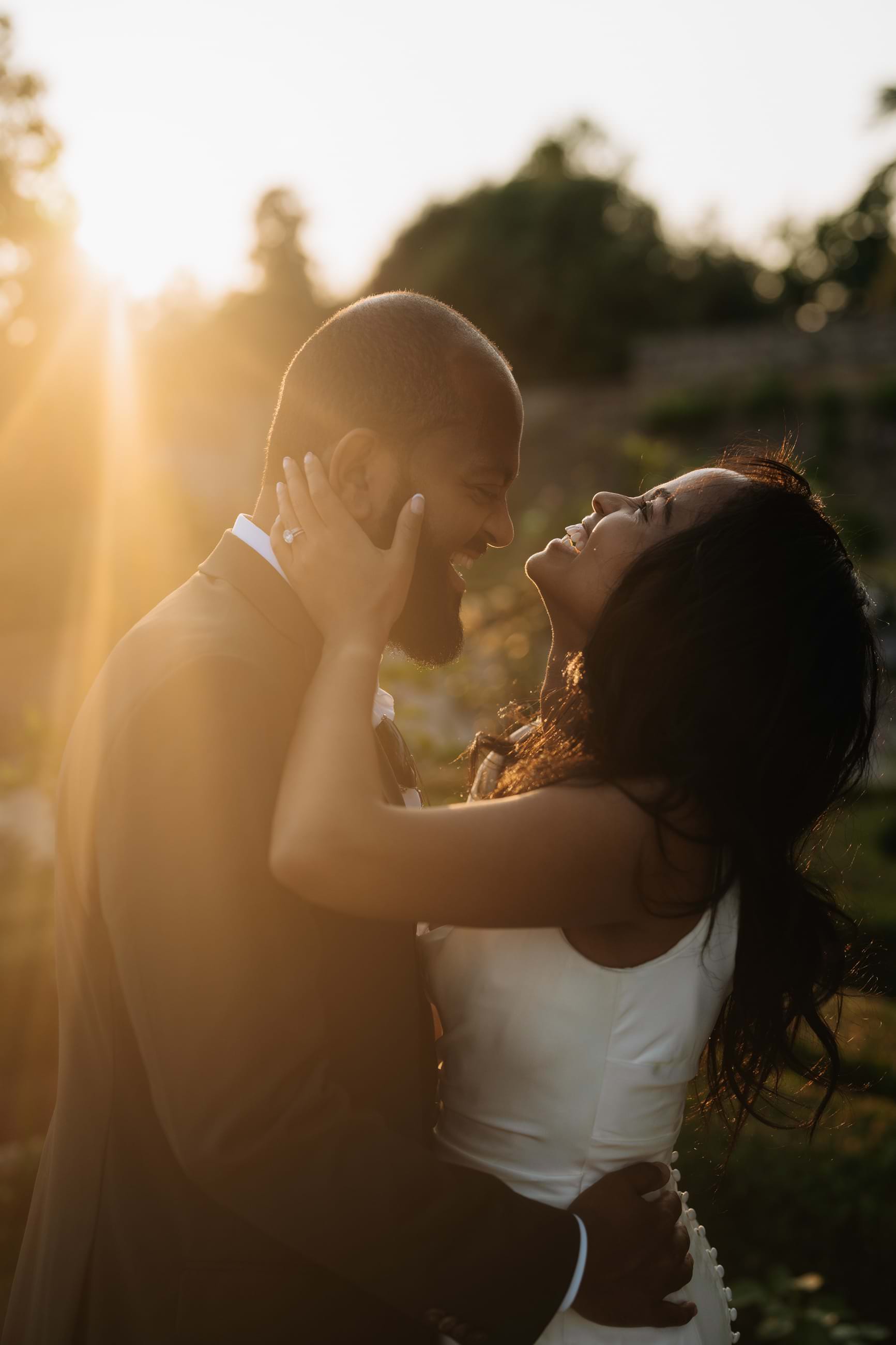 A couple embraces at sunset, bathed in warm, golden light. The woman in a white dress smiles up at the man in a dark suit, her hand gently on his face. They stand closely amid soft-focus nature, creating a romantic and intimate atmosphere—a timeless moment beyond packages and costs. Tuscan Wedding Photographer
