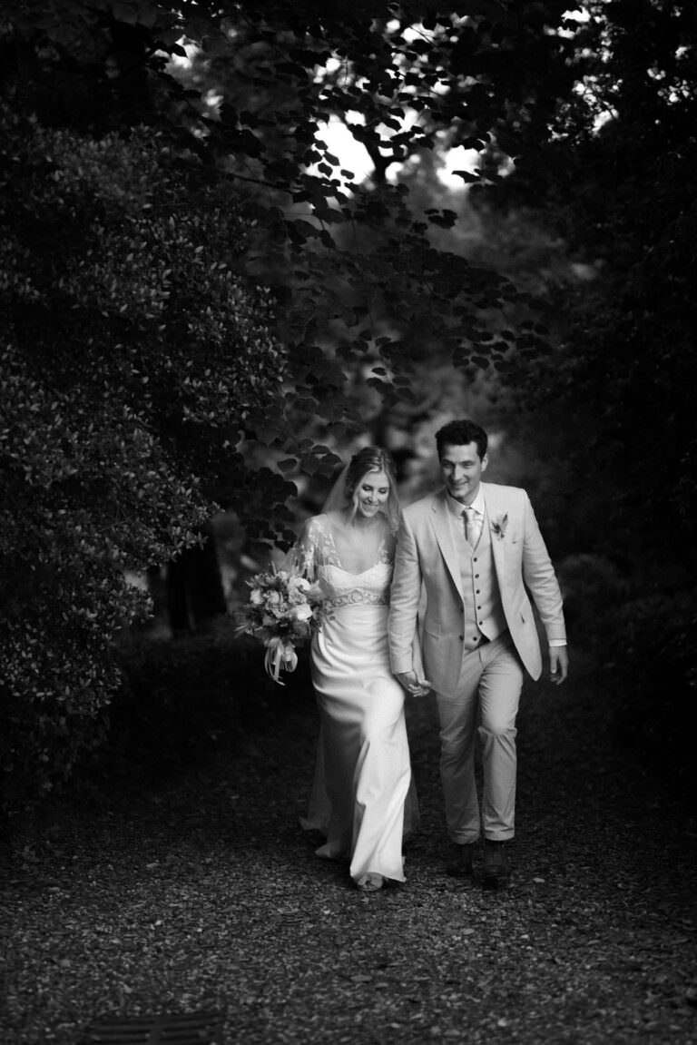 A bride and groom walk hand in hand down a tree-lined path at their British wedding at Monaciano. The bride wears a long, elegant dress, the groom in a light-colored suit. Both are smiling, surrounded by lush greenery in Federico Pannaccis serene black and white capture. Tuscan Wedding Photographer