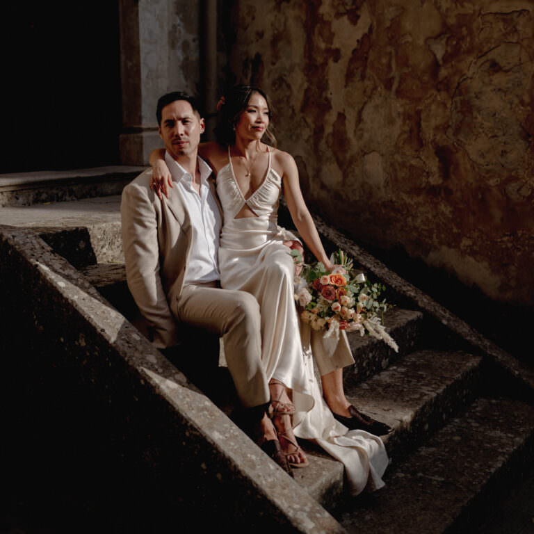 A bride in a flowing white dress holds a bouquet of orange and white flowers, sitting on stone steps beside a groom in a light-colored suit. Captured by an adept Sicily wedding photographer, the scene features an aged, textured wall. Soft lighting casts shadows, creating an intimate and romantic atmosphere. Tuscan Wedding Photographer