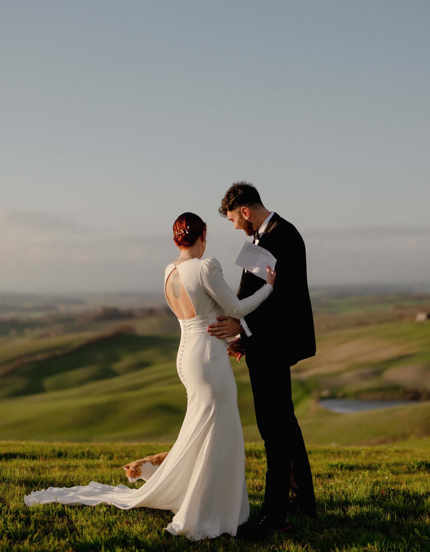 A bride in a long white gown and a groom in a black suit stand on a grassy Tuscan hilltop, facing each other. The brides dress has button details down the back. The groom holds papers. A small brown and white dog lays on the grass nearby, while rolling hills stretch under a clear sky—a perfect elopement scene. Tuscan Wedding Photographer
