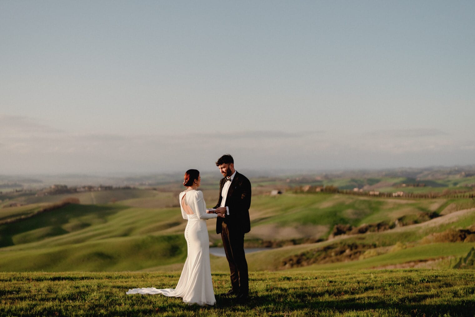 A couple stands outdoors on a lush, green hillside in Tuscany during sunset. The woman is in a white wedding dress, and the man is in a dark suit. They face each other, holding hands. The scene embodies the romance of eloping under an expansive sky with soft, warm lighting over rolling hills. Tuscan Wedding Photographer
