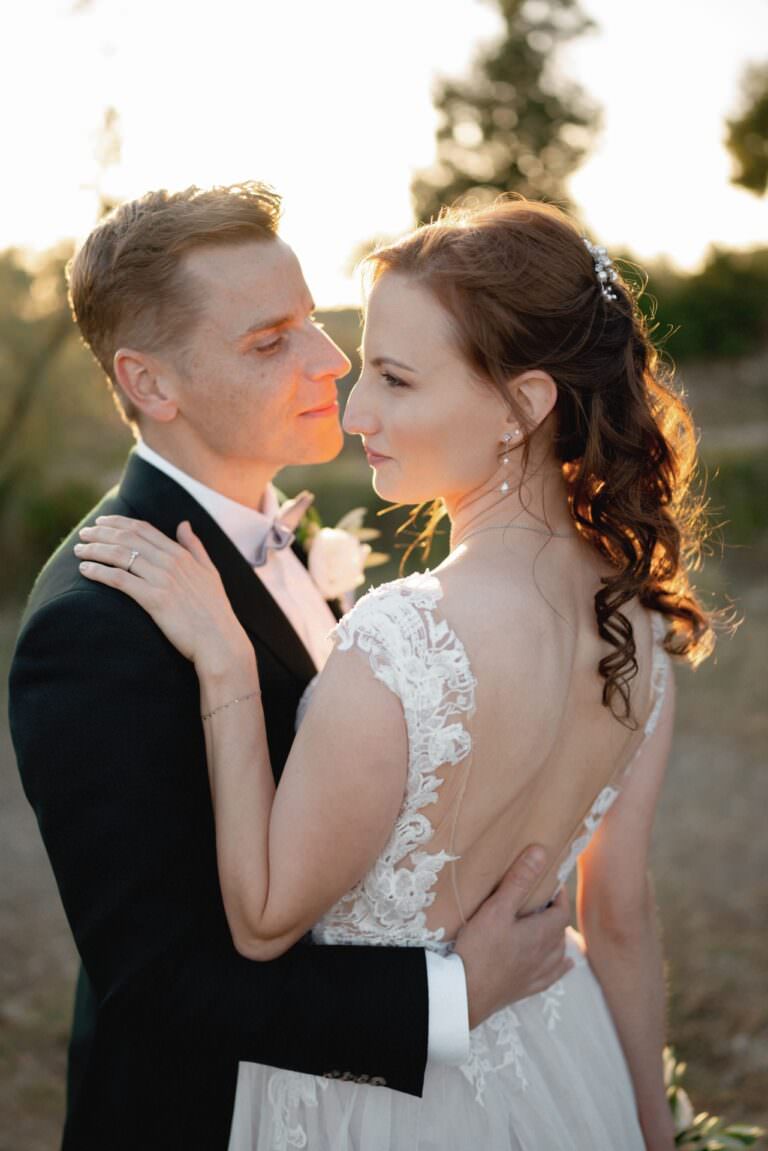 At Fattoria e Villa di Rignana in Tuscany, a couple embraces outdoors at sunset. The groom, in a black suit and bowtie, gazes lovingly at the bride in her lace wedding dress with an open back. Her curled hair is adorned with a decorative clip as warm sunlight bathes them in romance against a blurred natural backdrop. Tuscan Wedding Photographer