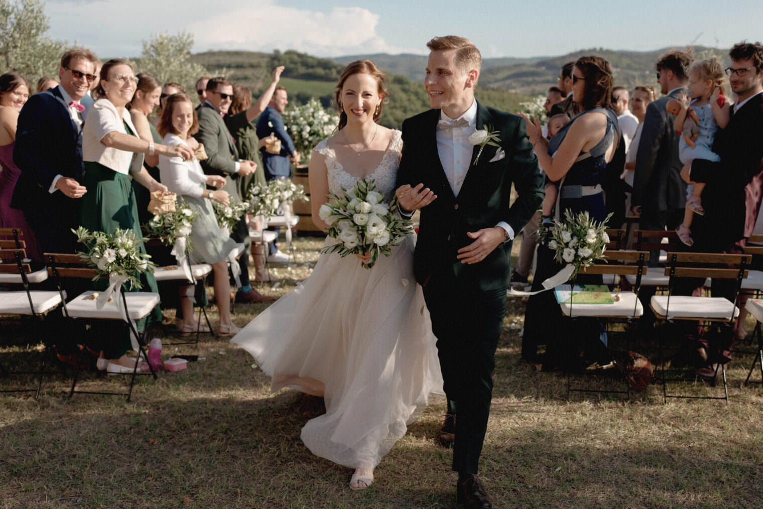 A joyful bride and groom, both smiling, walk down the aisle outdoors at Fattoria e Villa di Rignana in Tuscany. The bride holds a bouquet of white flowers and wears a flowing gown, while the groom is in a dark suit. Guests cheer amid scenic trees and hills, capturing this magical moment. Tuscan Wedding Photographer